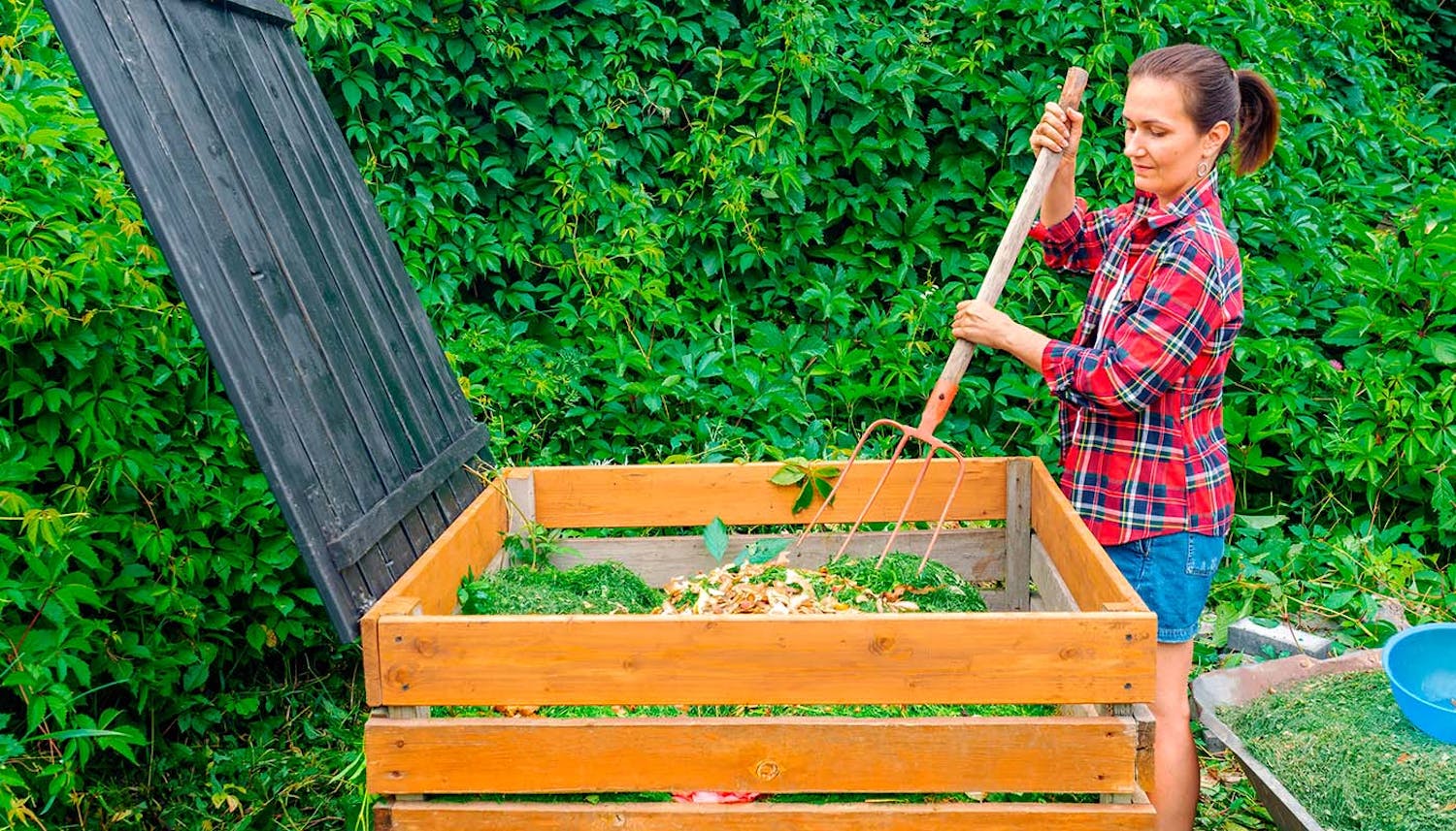 jordförbättring häck Kvinna använder en högaffel för att blanda kompost i en kompostbehållare av trä i en trädgård med gröna blad.